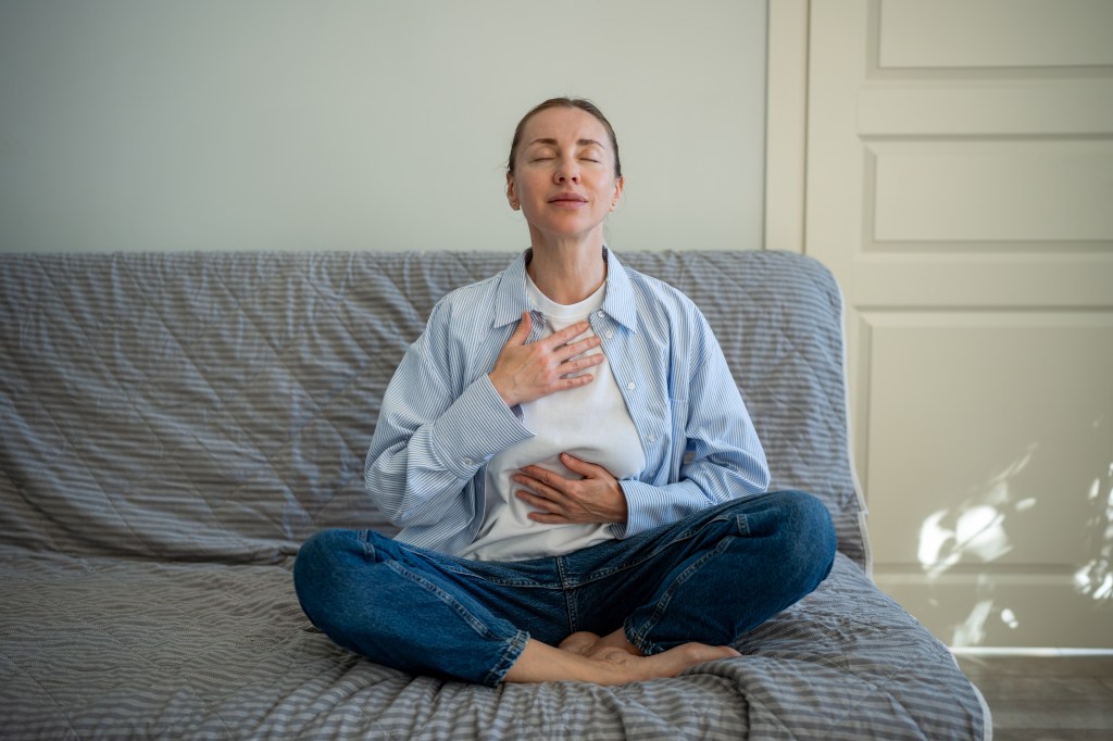 Woman in lotus pose with closed eyes, hands on chest and belly, practicing meditation at home.