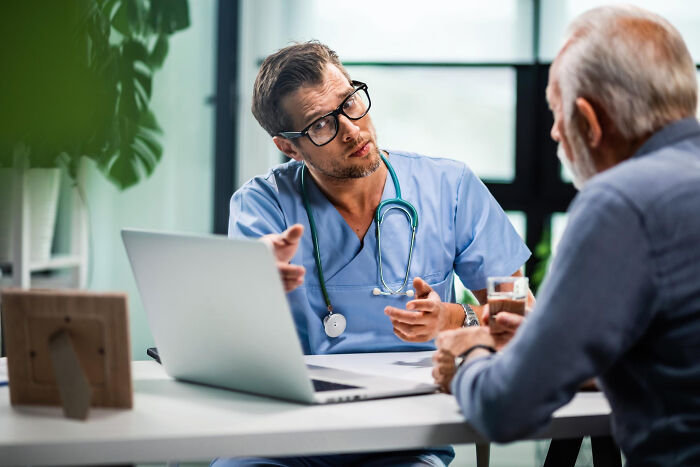 Doctor in blue scrubs speaking to an elderly patient while using a laptop, illustrating Dr Google versus real doctor concept.