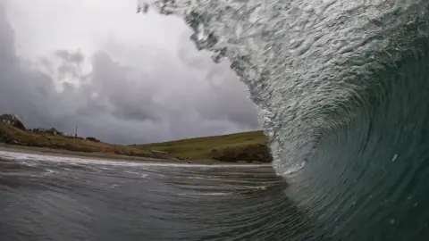 Emma Williamson A powerful ocean wave curls dramatically, forming a tunnel of foamy water near the shore. The coastline in the background features grassy hills and a few scattered buildings under a stormy, grey sky.