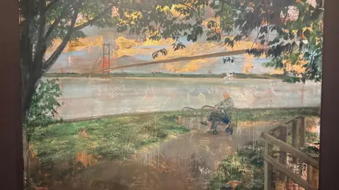 Emma Prempeh A photo of a painting hanging in a gallery, the paining portrays a man sat resting on a walking aid looking out towards the River Humber, with the Humber Bridge behind him. A tree frames the scene.