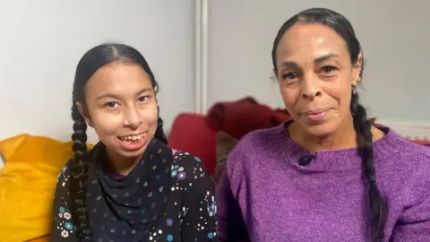 Phil Shepka/BBC Neave and Natasha are sitting indoors on a sofa. Both have long, dark hair styled in braids/pigtails. Natasha, who is taller, is wearing a purple knitted top with a small microphone clipped near the neckline. Naeve is dressed in a black top patterned with colourful stars and flowers. They are both looking at the camera and smiling. 