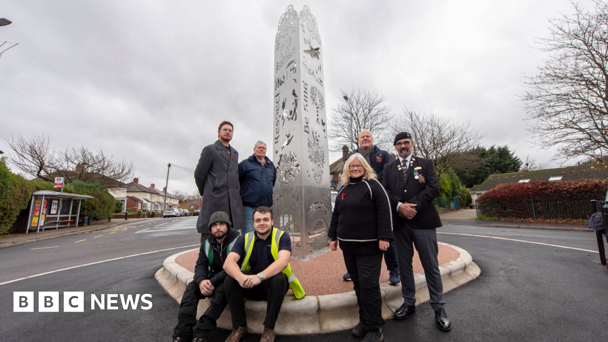 A group of seven people are in front of a metal sculpture, which looks to be in the middle of a roundabout. On the left of the sculpture two of the men are sitting, while two others stand behind them. To the right two men and one woman are all standing