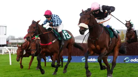 PA Media Two jockeys are racing their horses either side of eachother, and behind them is a hurdle. They are racing on grass.