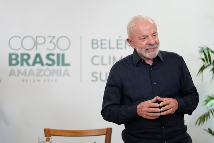 an older man with white hair and beard wearing black clothes stands in front of a sign reading COP30 Brasil