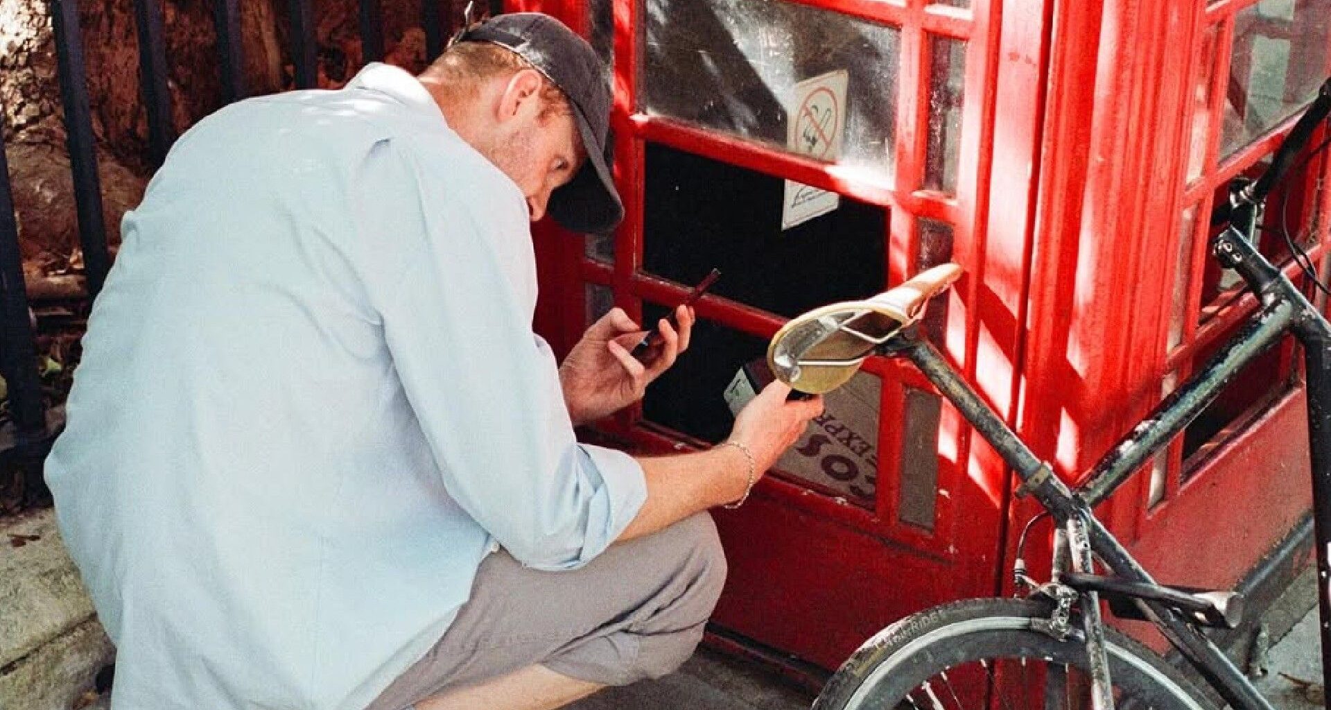 A man wearing a light blue shirt and a baseball cap is crouching down next to a red telephone booth, appearing to be examining something inside or on the ground while holding a small black object and a bottle.