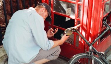 A man wearing a light blue shirt and a baseball cap is crouching down next to a red telephone booth, appearing to be examining something inside or on the ground while holding a small black object and a bottle.