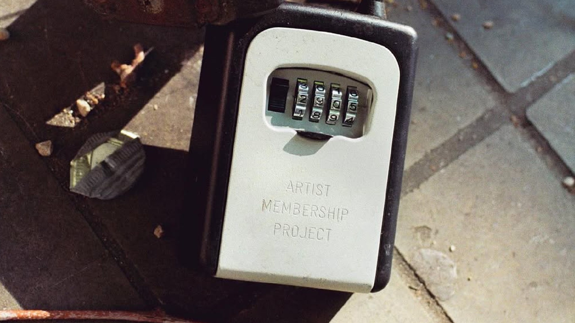A close-up shot of a key safe box on the ground with a four-digit combination lock, inscribed with the text "ARTIST MEMBERSHIP PROJECT" on its pale gray face.