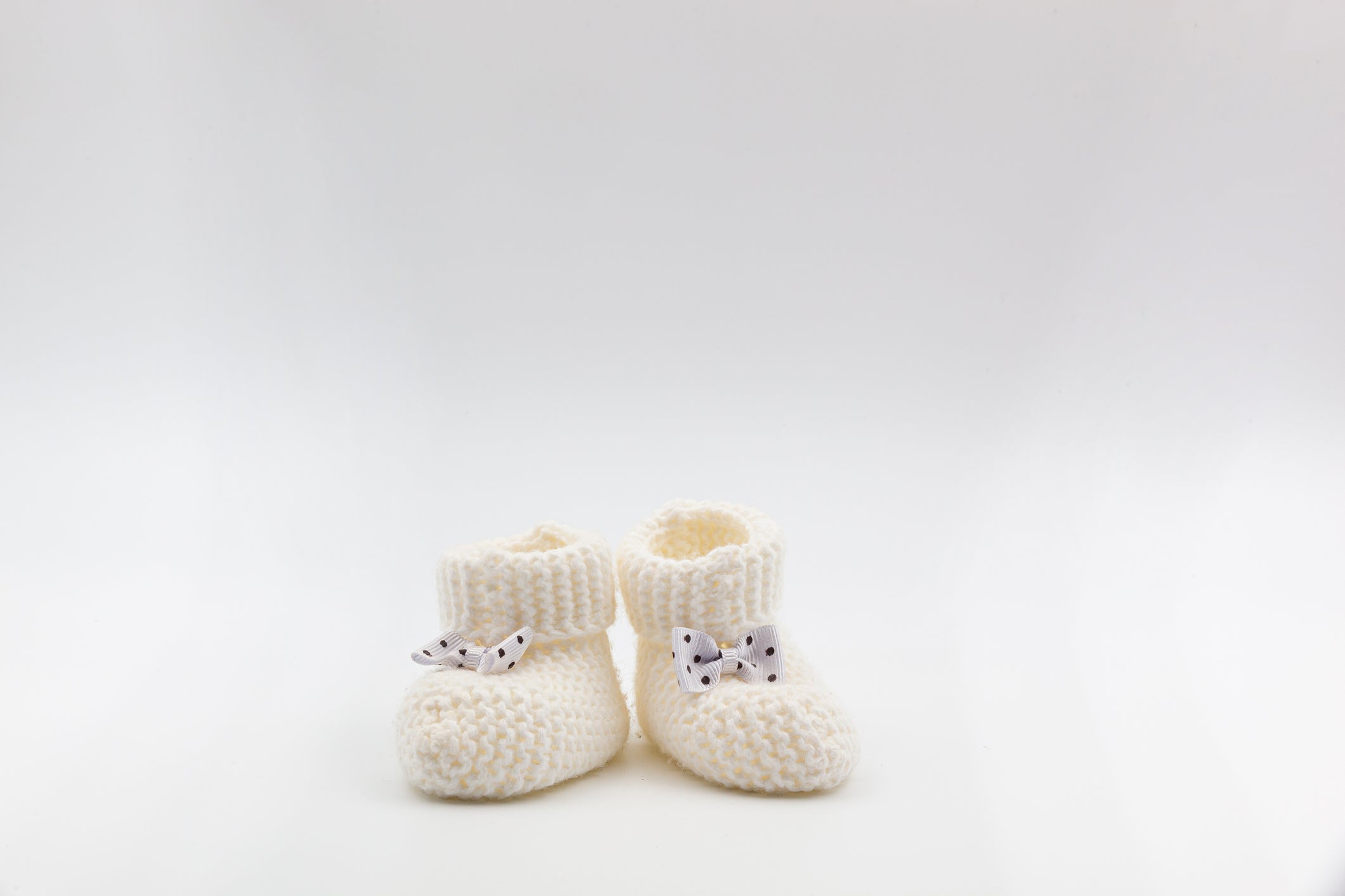 Knitted white baby booties with polka-dot bows on a gray isolated background.