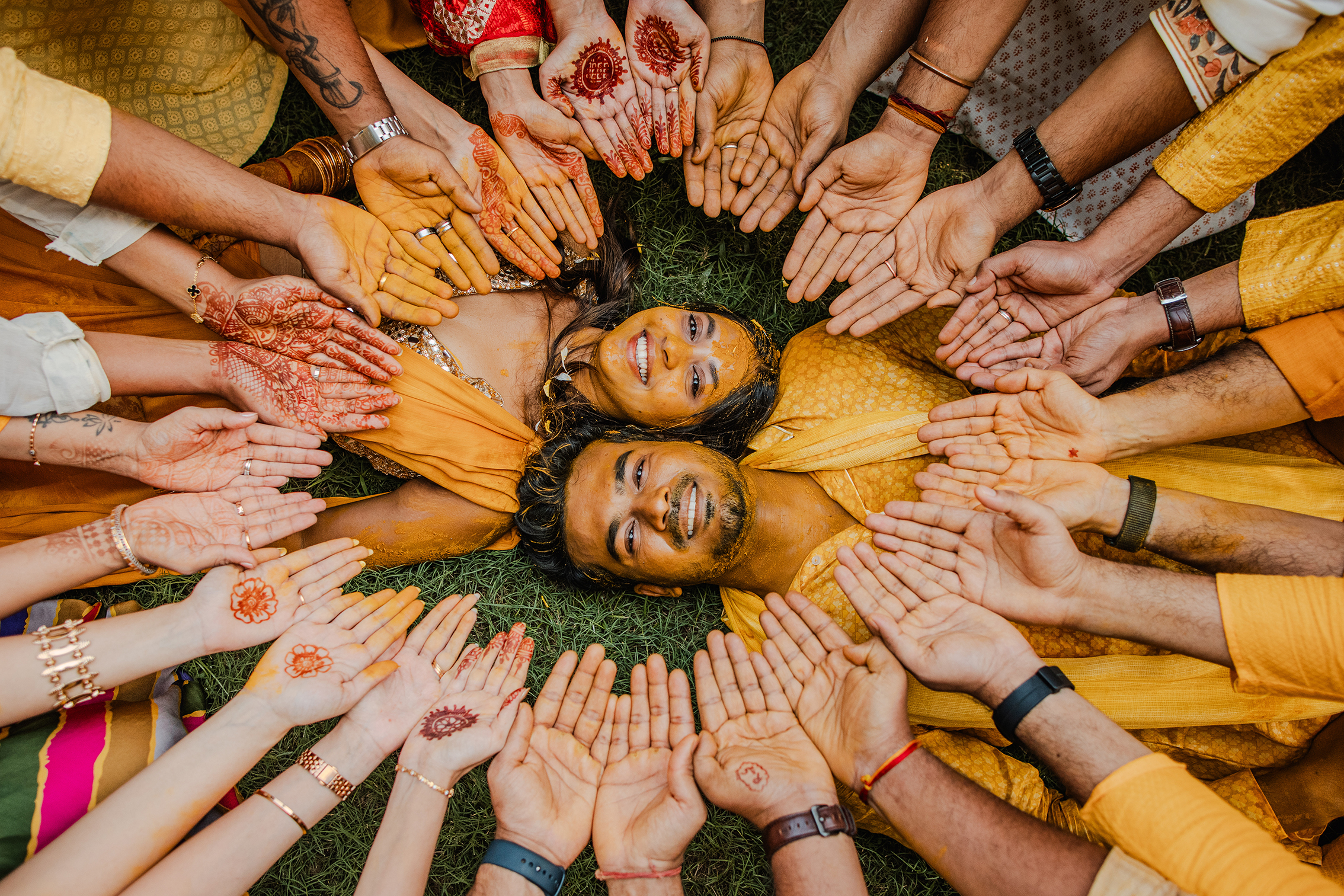 A joyful couple lies on grass surrounded by outstretched hands decorated with henna. The scene is vibrant and celebratory, conveying cultural festivity