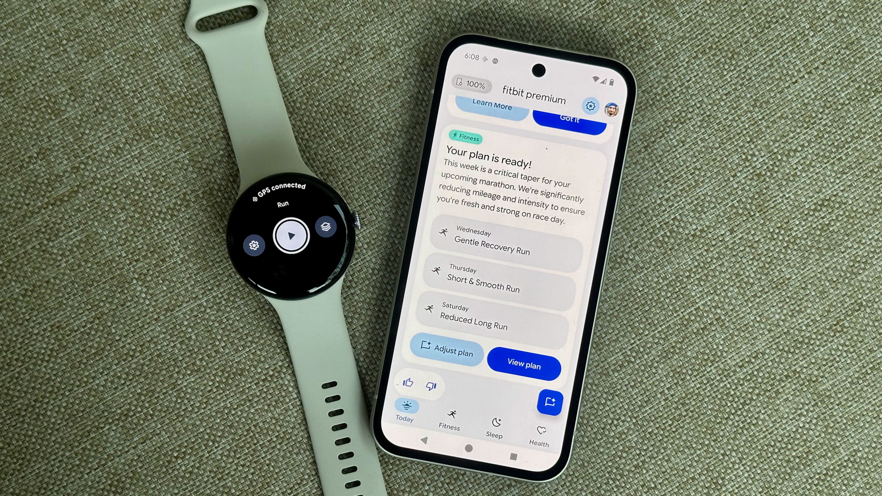 A photo of the Google Pixel Watch 4 sitting next to a Pixel 9a open to the Fitbit app, showing the Fitbit Premium Health Coach preview with the Fitness tab open; it shows a customized workout plan with three upcoming runs.