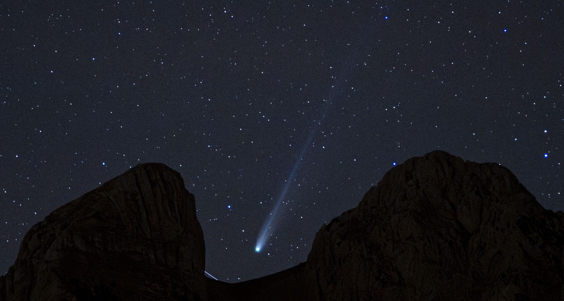 A white streak of a comet's tail can be seen in the center of the image as it streaks diagonally from top right to lower left between the dark silhouette of rocks blocking part of the starry night sky