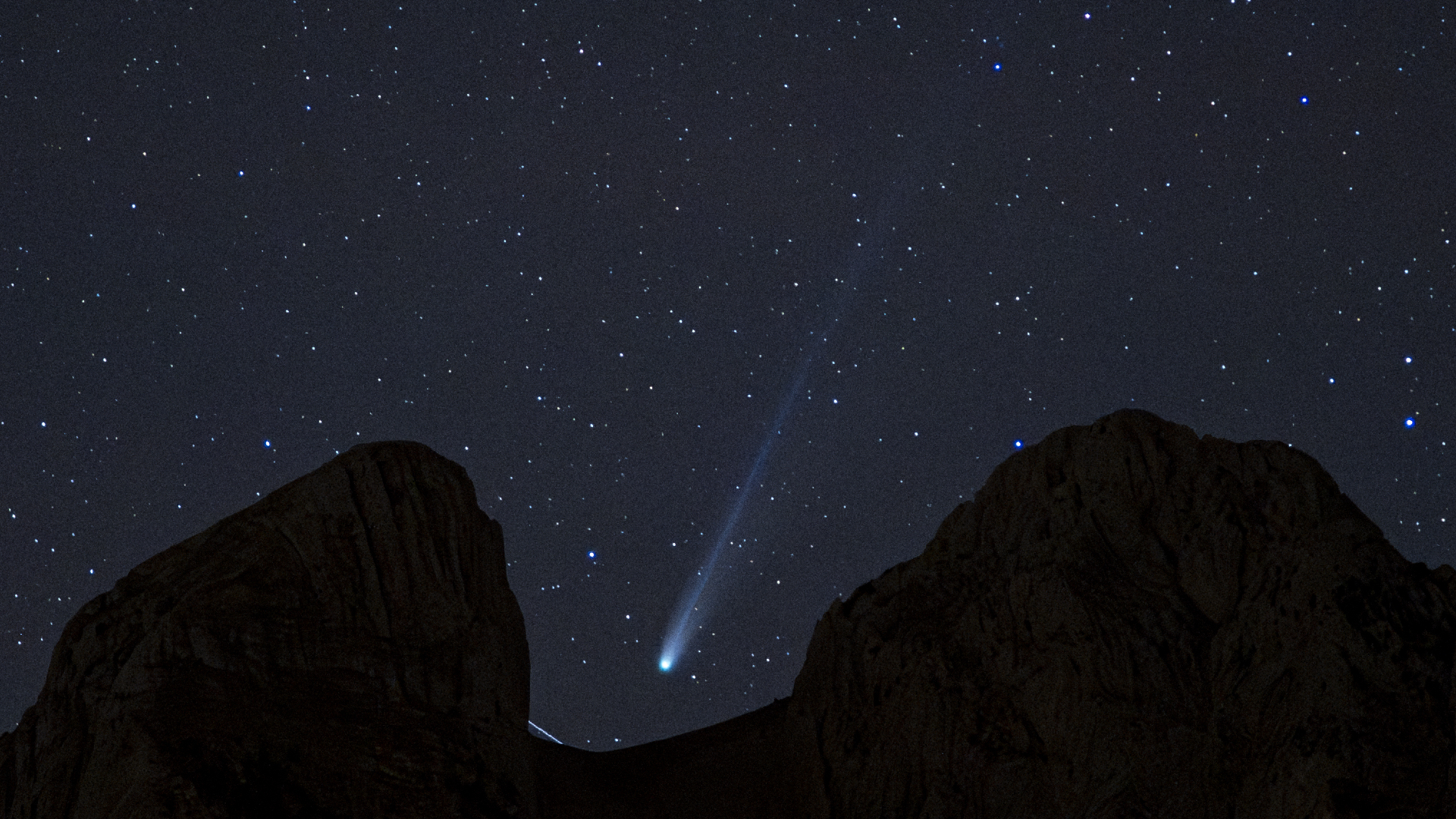 A white streak of a comet's tail can be seen in the center of the image as it streaks diagonally from top right to lower left between the dark silhouette of rocks blocking part of the starry night sky