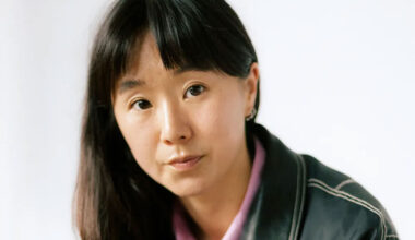 a woman with long dark hair and bangs seated with her elbows on her knees and her hands clasped, wearing a black leather jacket and a pink button down underneath, with a serious expression