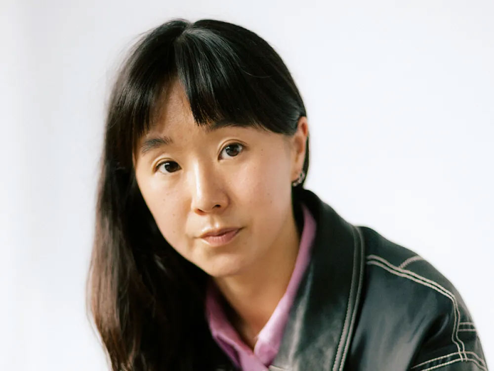 a woman with long dark hair and bangs seated with her elbows on her knees and her hands clasped, wearing a black leather jacket and a pink button down underneath, with a serious expression