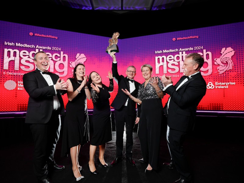 A group of people dressed up in black tie cheering on stage holding an award. A screen behind them says Medtech Rising.