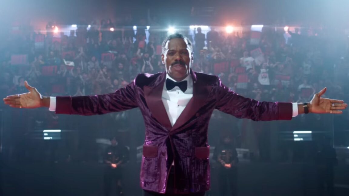 black man in colorful purple suit with arms outstretched in front of a camera with huge crowd behind him