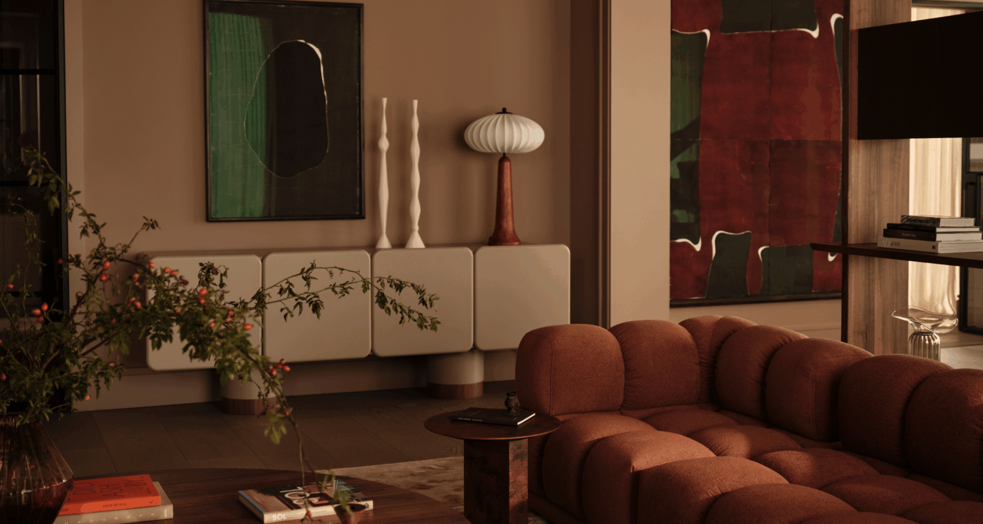 A modern living room with a contemporary rust sofa, earth tones, decorative accessories and artwork and a vase of foliage with berries on the round coffee table