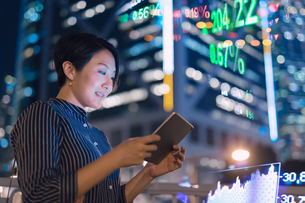 Investor reviewing figures on tablet while standing opposite a building displaying stock numbers.