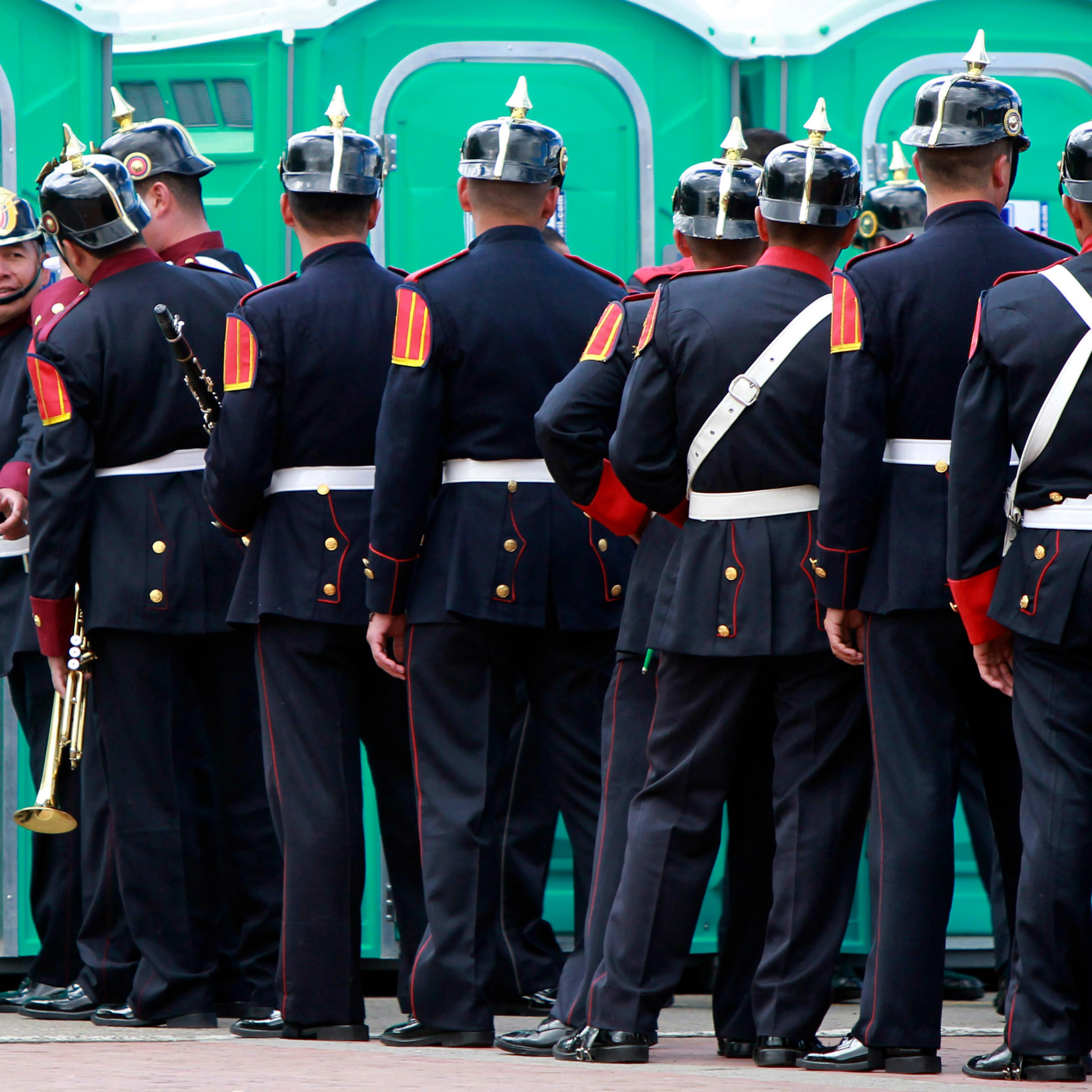 Photo of uniformed men standing outside portable toilets, one gestures invitingly.