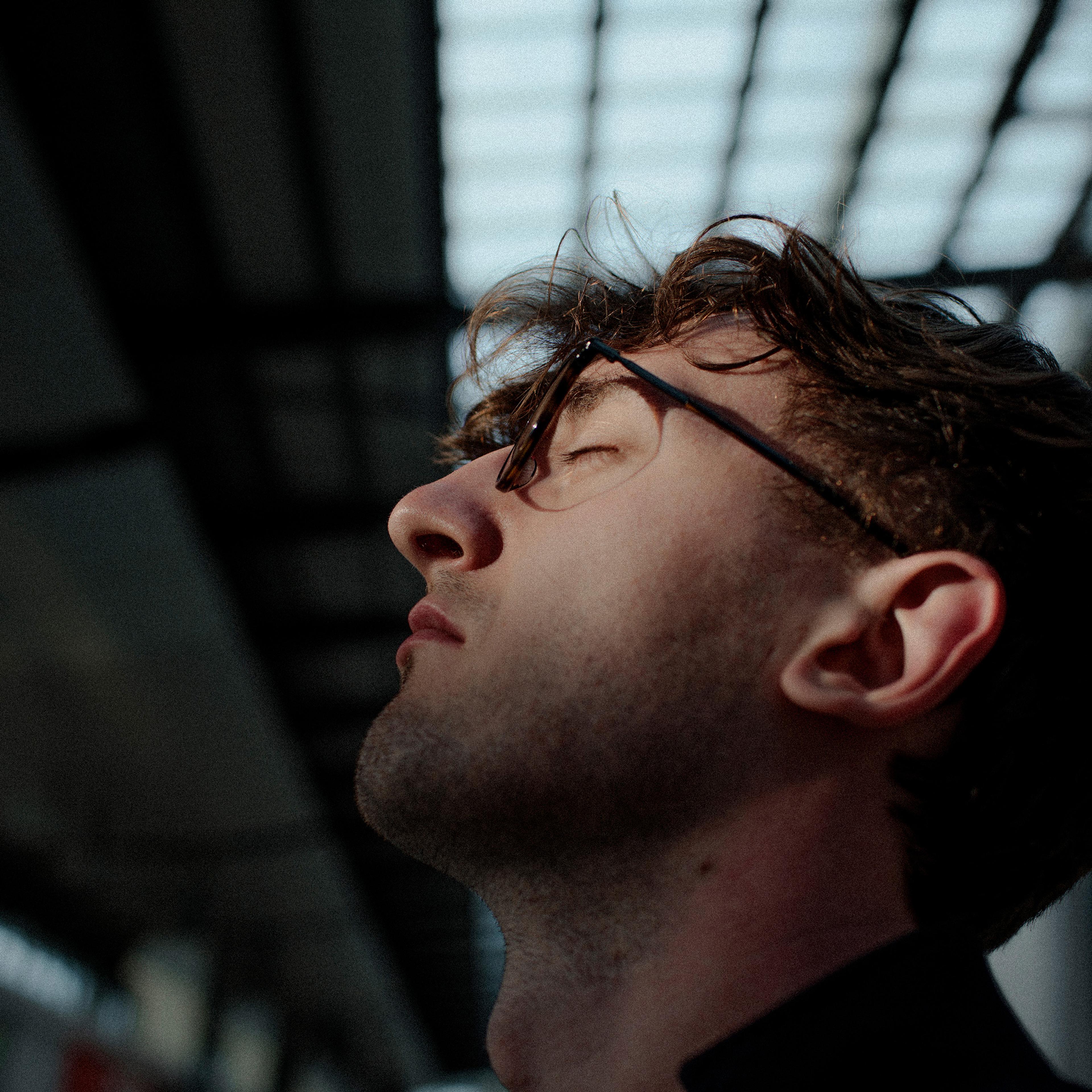 Photo of a man in profile with glasses, eyes closed, head tilted back under a structure with a grid-like roof.
