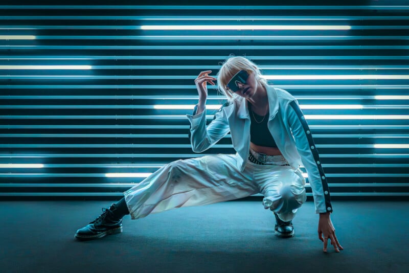 A person in white clothing and black boots poses in a dynamic, crouched stance in front of horizontal blue neon lights, wearing sunglasses and striking a futuristic, stylish look.