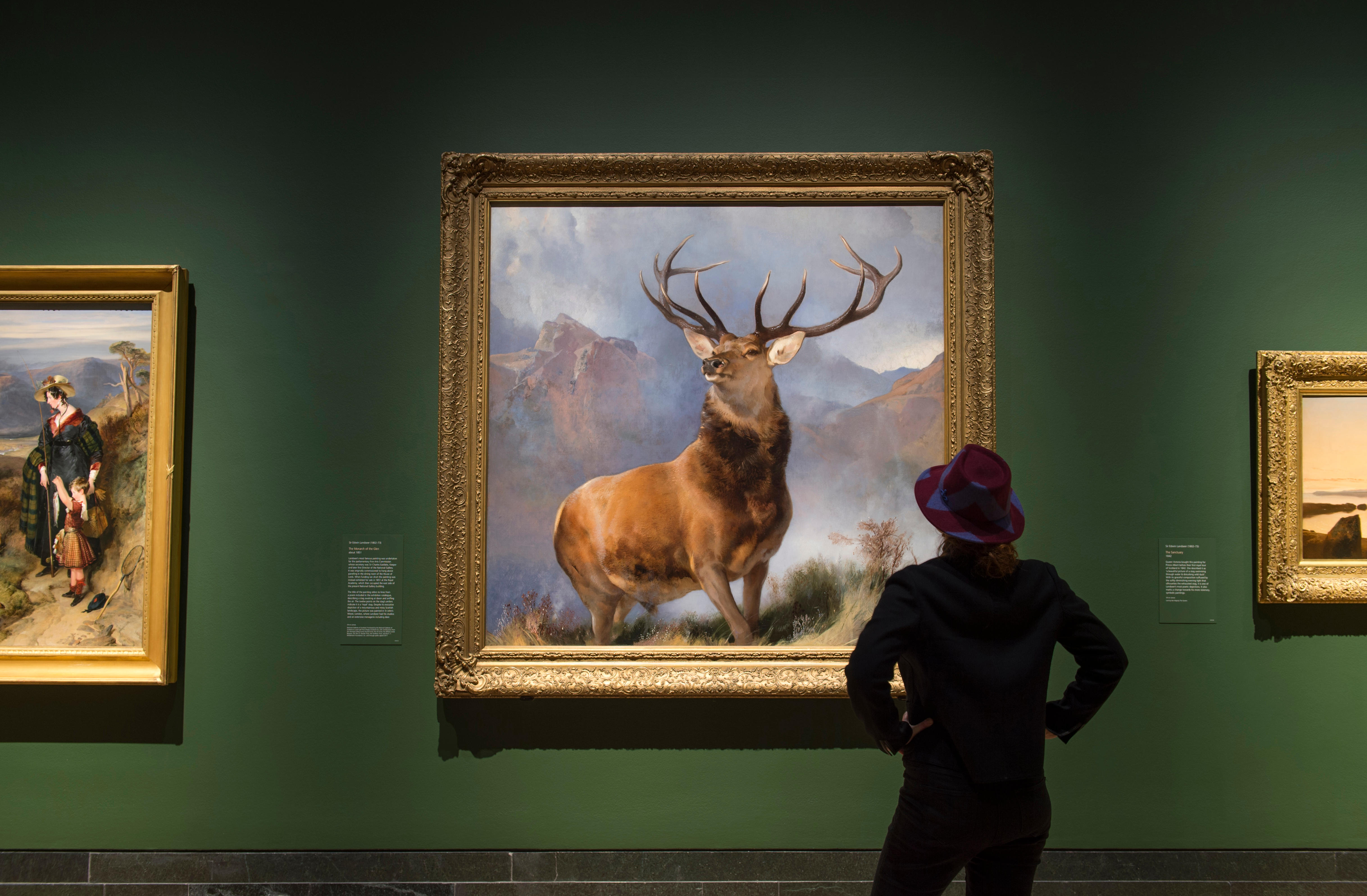 A person in a red and blue hat views the painting "The Monarch of the Glen" by Edwin Landseer at an exhibition.