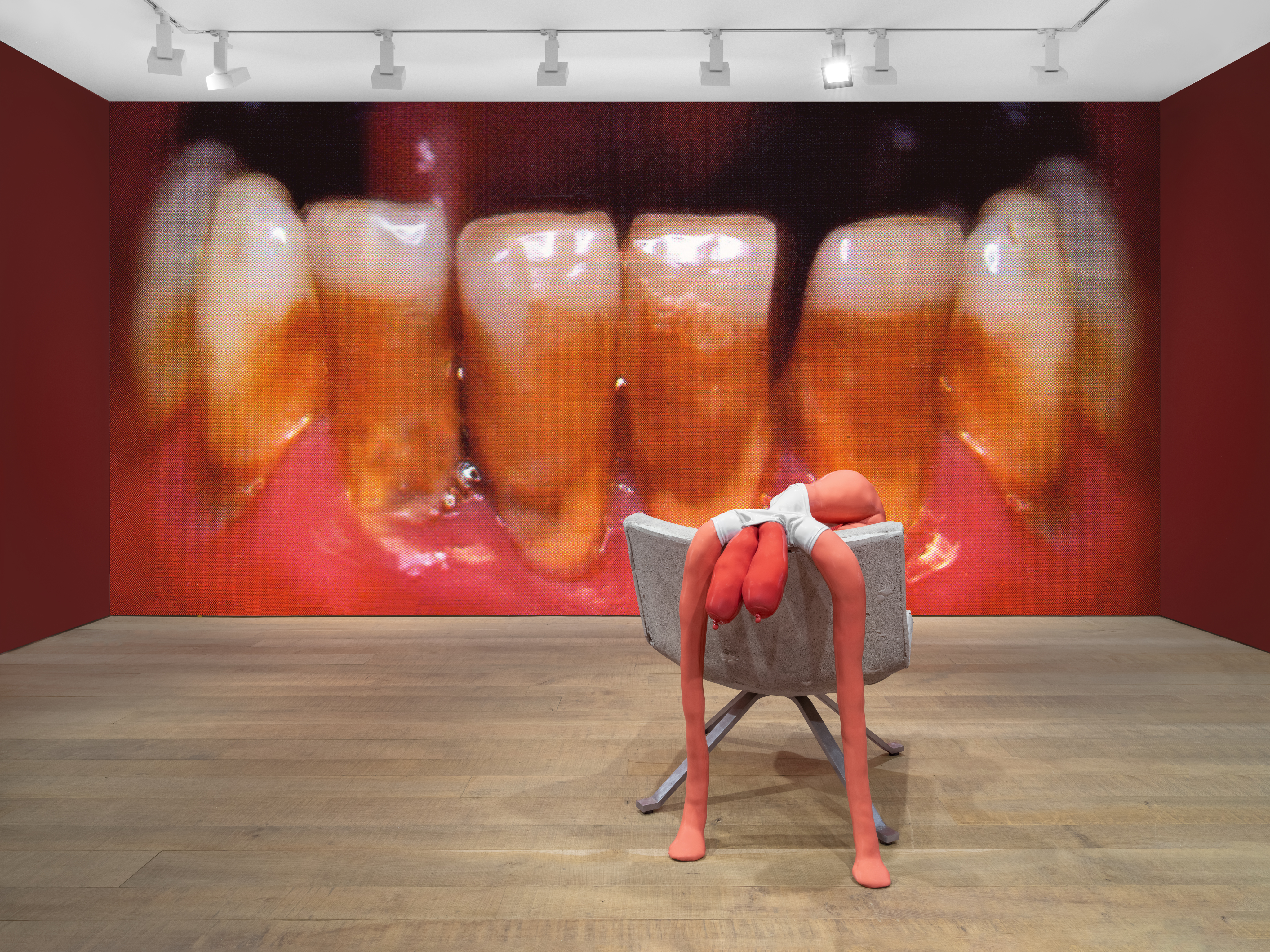 Exhibition view with a close-up of teeth on the wall and a pink, headless mannequin in a chair.