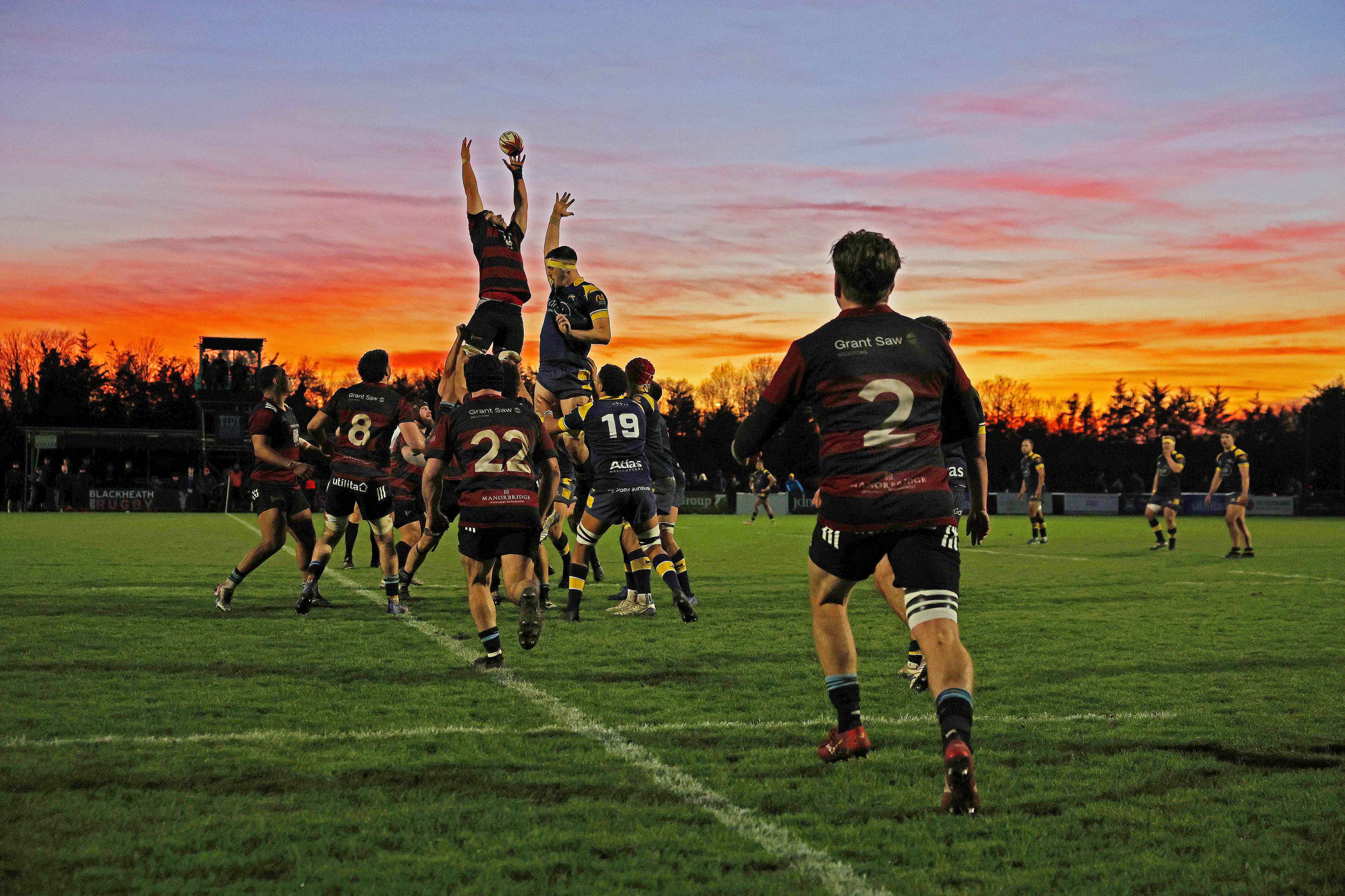 Rugby players from Blackheath and Leeds Tykes in action during a lineout against an orange sunset.