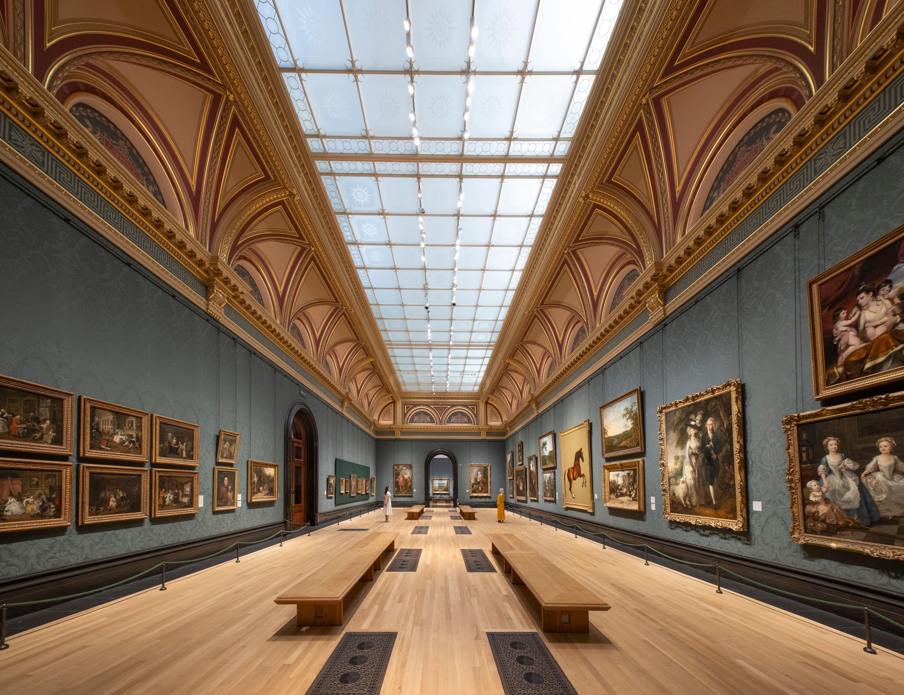 A grand hall in the National Gallery in London, featuring rows of classical paintings on teal walls, a gilded and vaulted ceiling, and a light wooden floor with long wooden benches in the middle. Two people are visible in the background looking at the art.