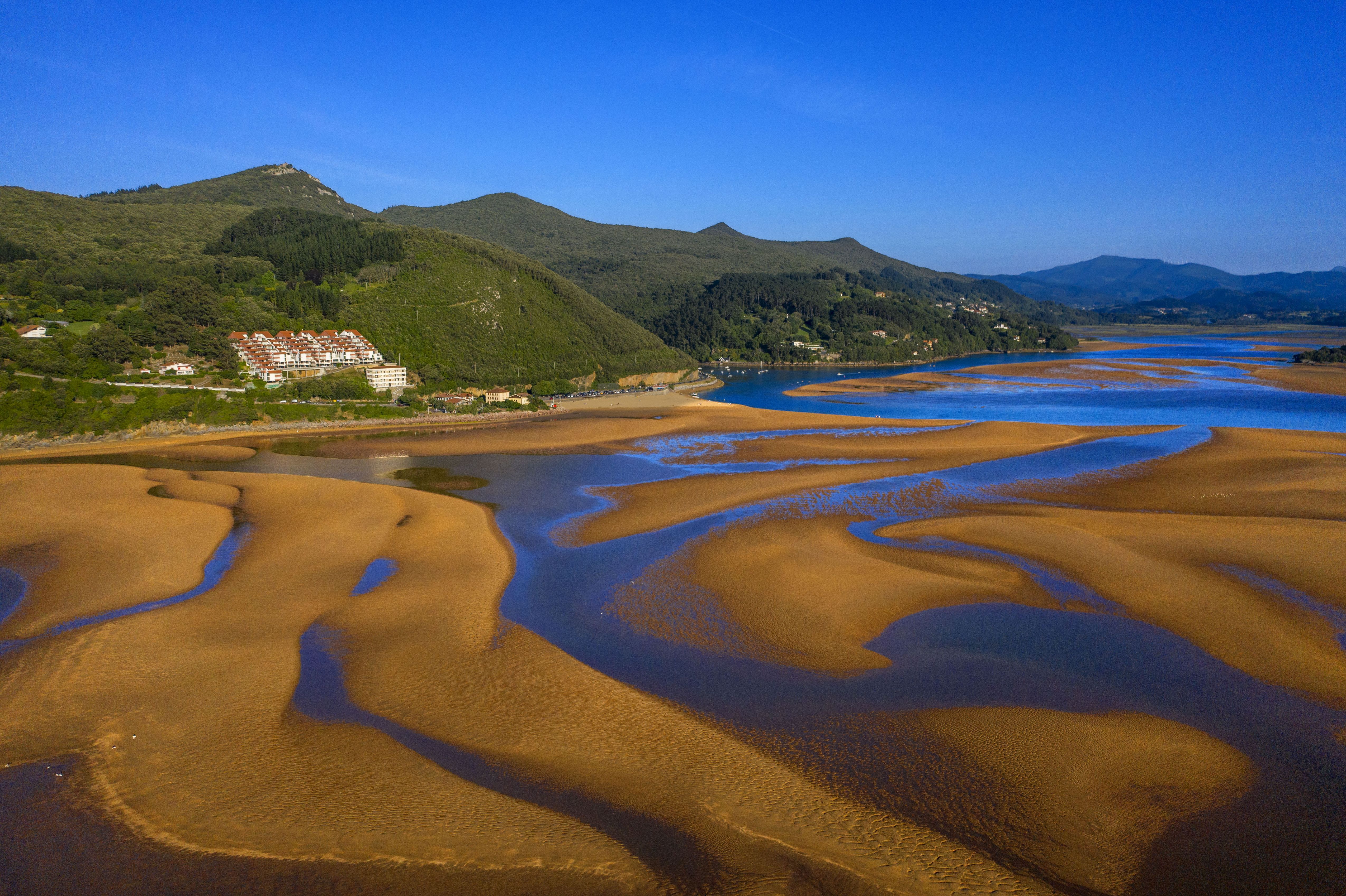 Aerial view of the Gernika estuary, Urdaibai Biosphere Reserve, showing sandbanks and blue water against a backdrop of green, forested hills with buildings.