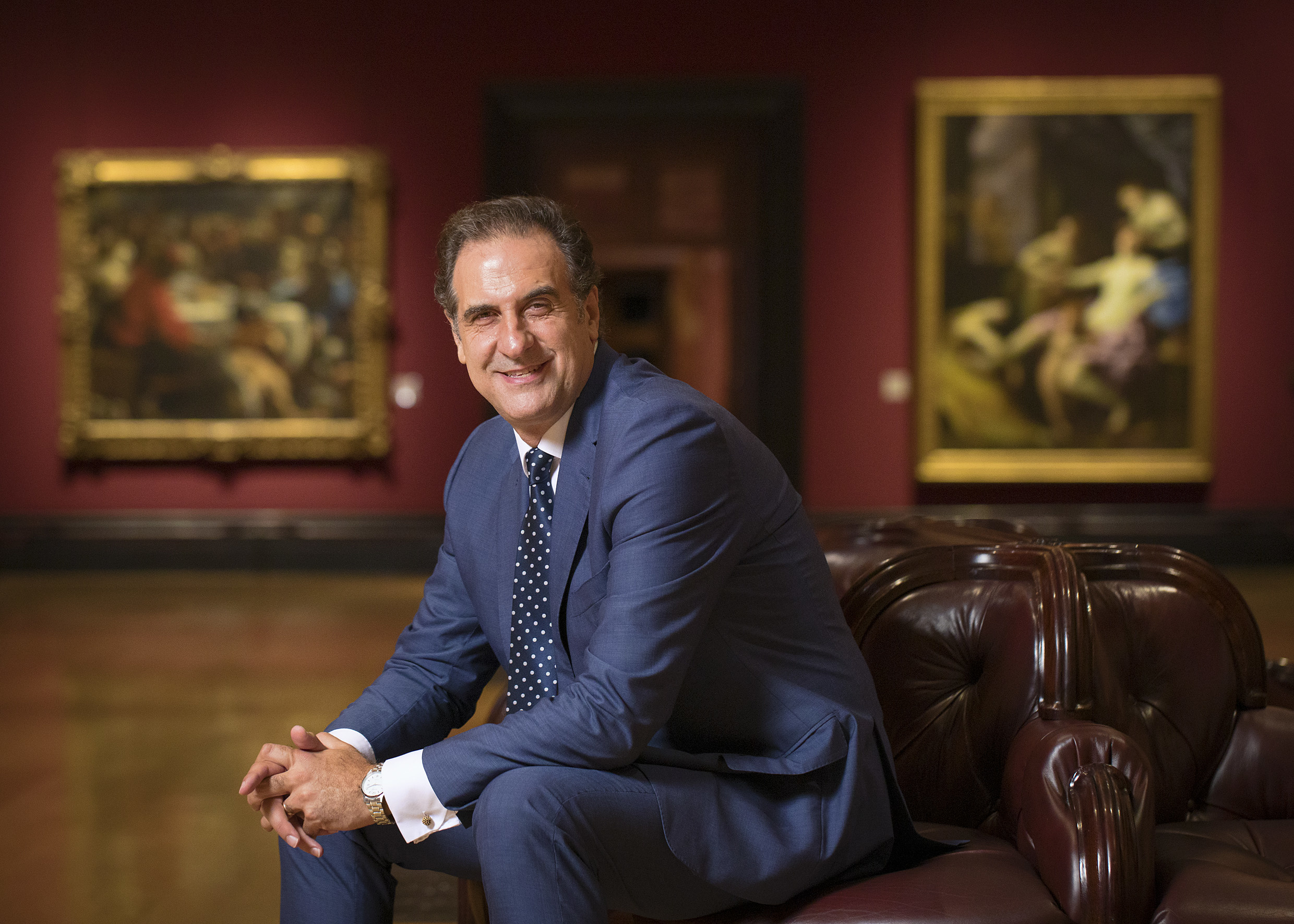 Gabriele Finaldi in a suit, sitting on a leather sofa in a gallery with paintings on the wall.