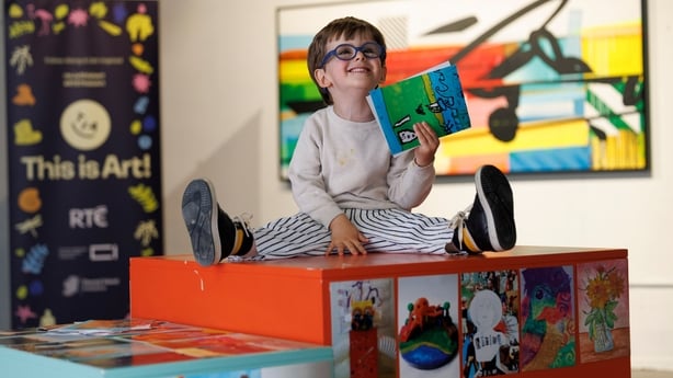 A young oy with glasses and a white jumper sits on an art display unti holding a postcard.