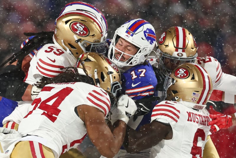 Football players in red and gold San Francisco 49ers uniforms tackle a Buffalo Bills player wearing a blue jersey with number 17 during a snowy game. Multiple players are colliding in intense action.