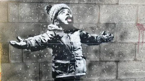 Banksy A lack and white mural of a small boy wearing a woolly hat, looking up with his mouth open, catching snowflakes
