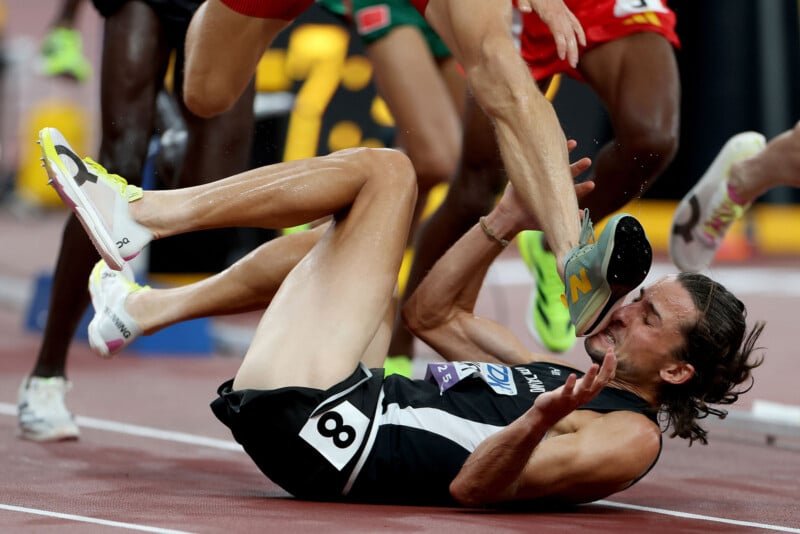 A runner in a black and white uniform falls backward onto the track as another athlete’s foot lands on his face during a race, with other runners’ legs visible in the background.