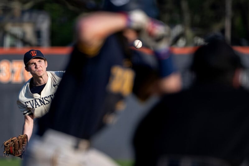 A baseball pitcher in an Evanston jersey throws a pitch towards a batter, who prepares to swing. The ball is mid-air, and an umpire is visible in the background, watching the play closely.