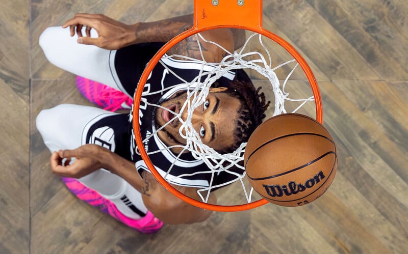 A basketball player in a black and white uniform looks up as a basketball passes through the hoop, viewed from above the rim on a wooden court.