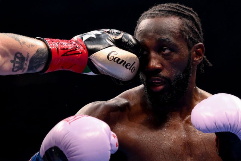 A boxer wearing white gloves labeled "Canelo" punches another boxer with braided hair and a focused expression in the face during a match. The impact distorts the recipient’s cheek and eye area.