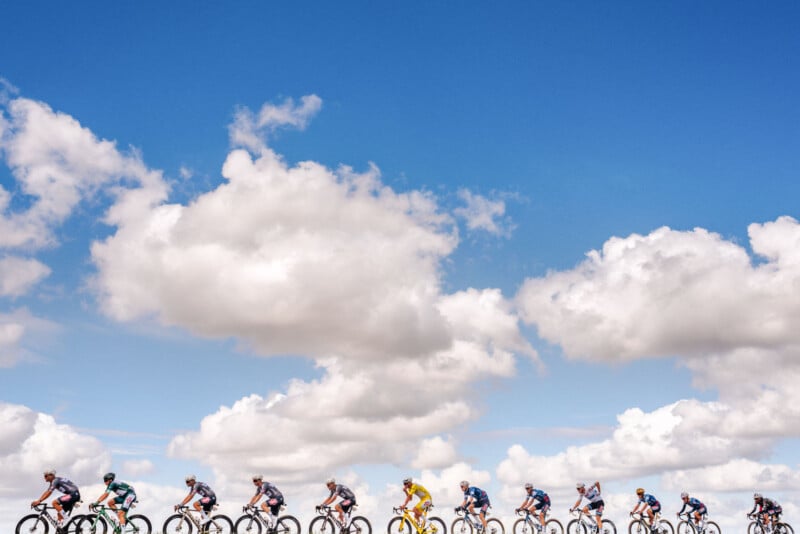 A group of cyclists rides in a line under a bright blue sky filled with large, fluffy white clouds. The scene is vibrant and energetic, set against the expansive sky.
