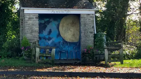 A bus stop flanked by benches and flowers is painted inside with a large harvest moon and a merging of different colour blues for the sky 