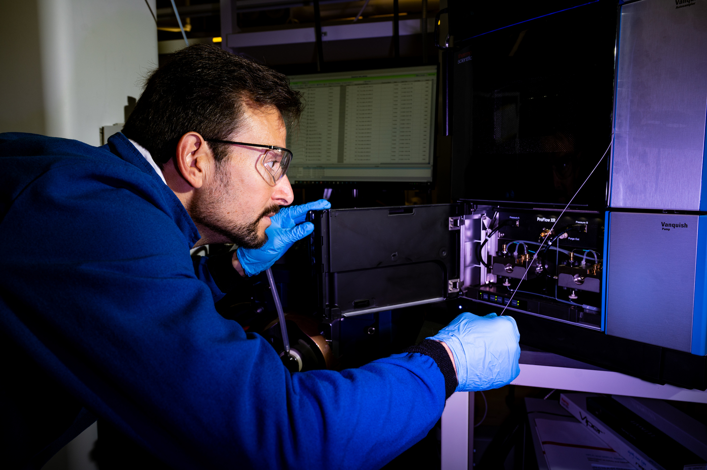 A man with glasses dressed in a blue lab coat tinkers with an electric panel in a lab.