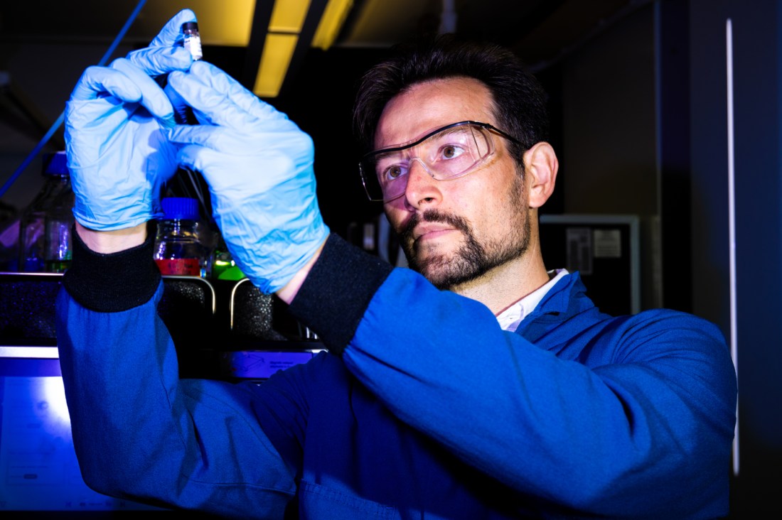 A scientist wearing blue gloves and safety glasses holds up a small vial to examine it under bright lab lights, surrounded by equipment and bottles in a research laboratory.