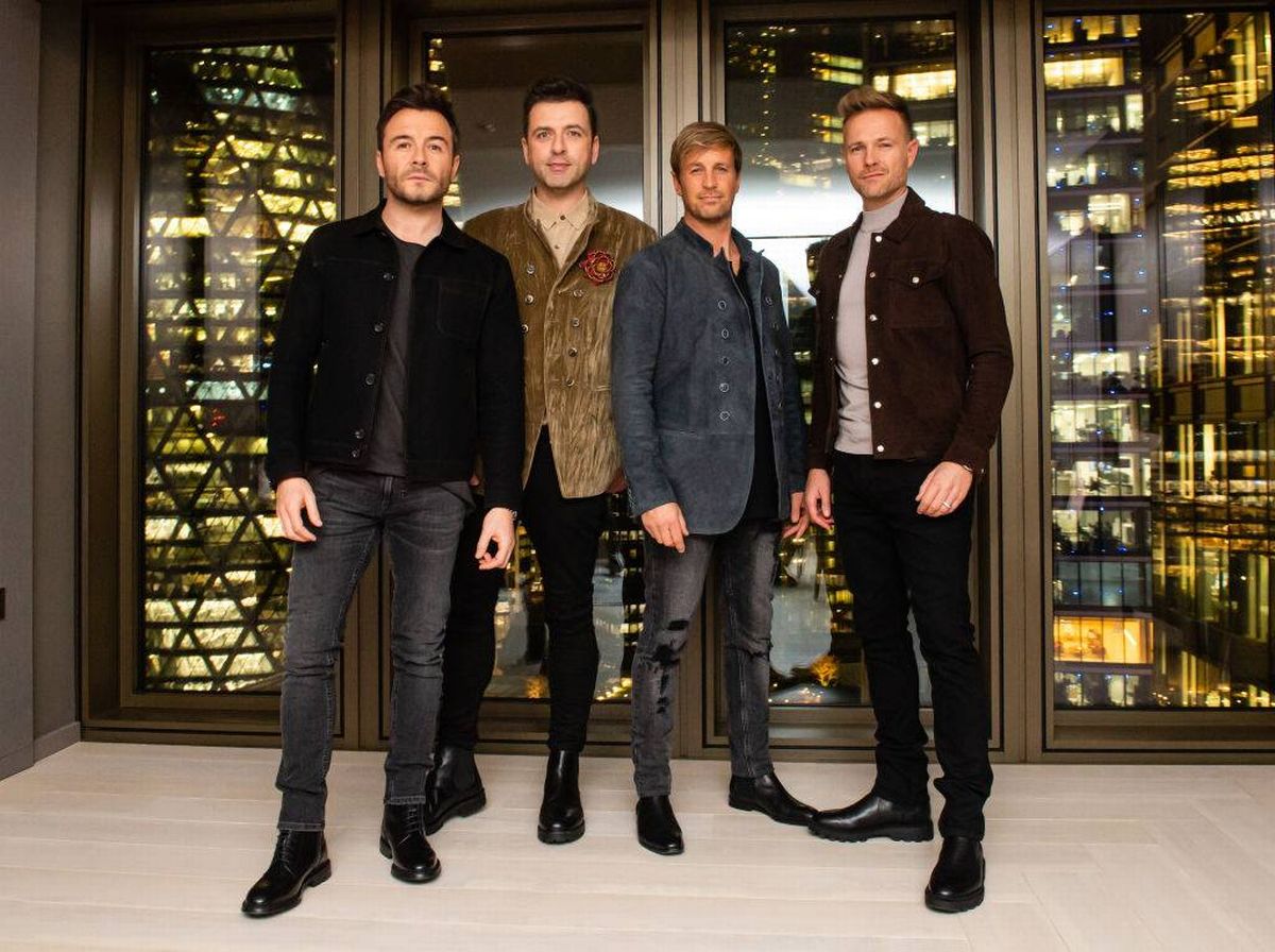 Three individuals dressed in formal attire stand side by side in front of an architectural glass structure with illuminated cityscape reflections in the background.