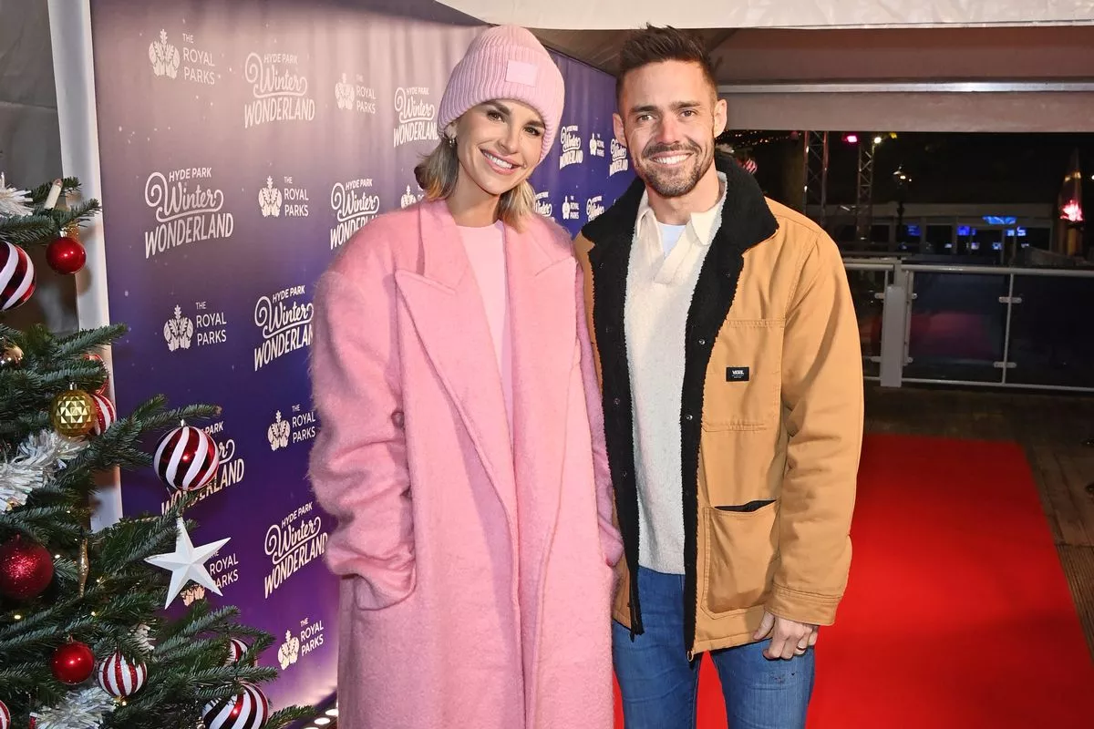 Two individuals dressed warmly are standing side by side on a red carpet, adjacent to a festively decorated Christmas tree.
