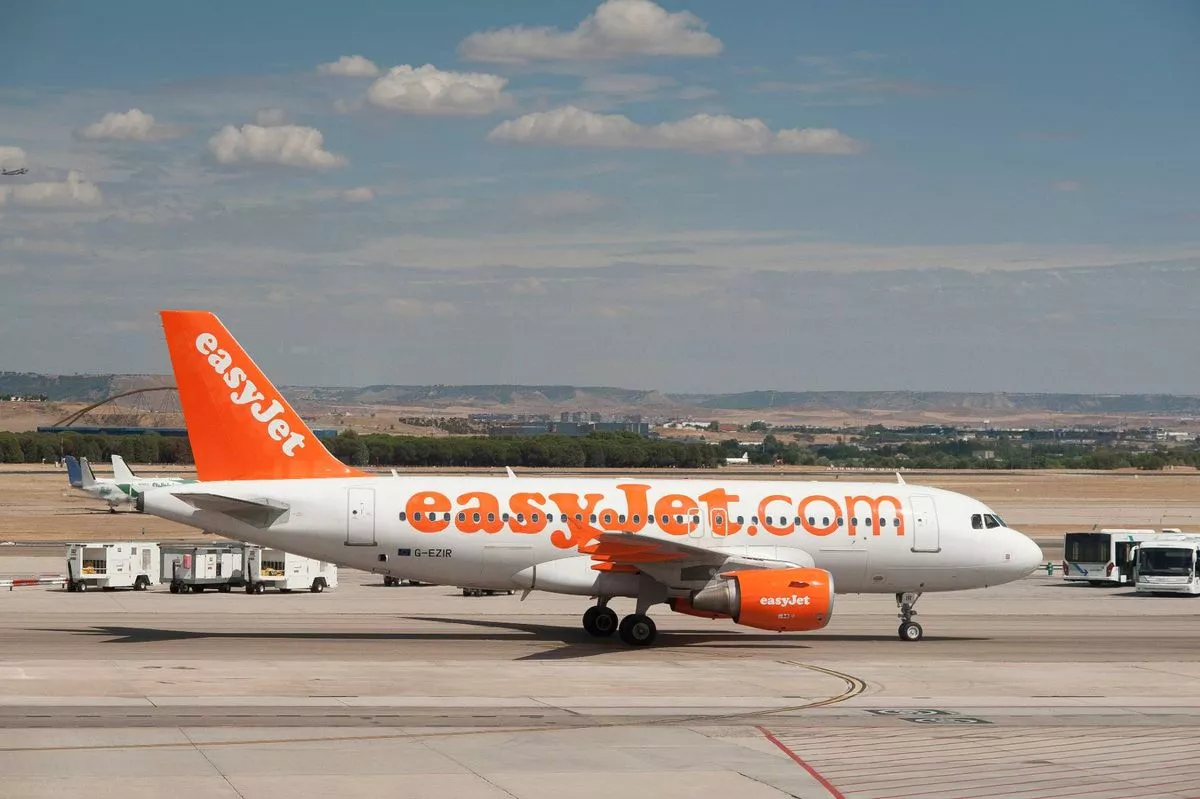 An orange and white commercial airplane, identified as an easyJet aircraft, is stationed on the runway of an airport, with a clear blue sky and scattered clouds in the background.