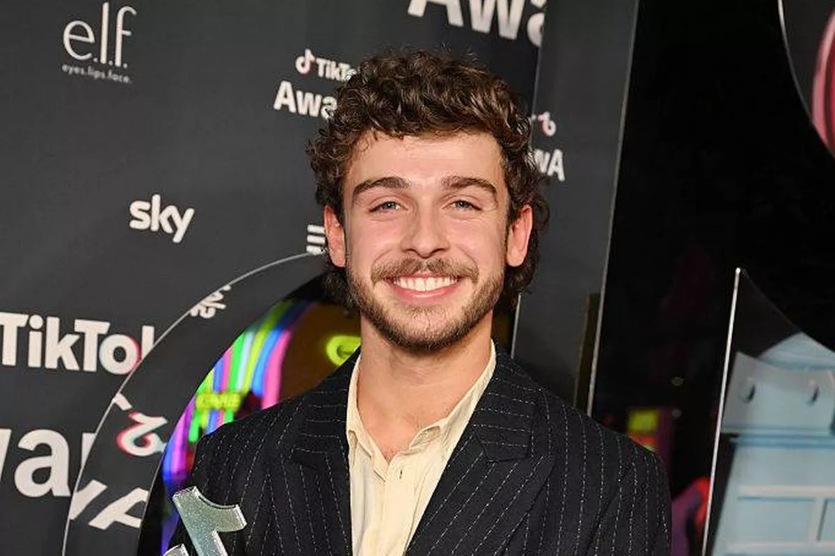 An individual dressed in a dark suit, standing in front of a backdrop adorned with logos and text, is holding a trophy and smiling for the camera.
