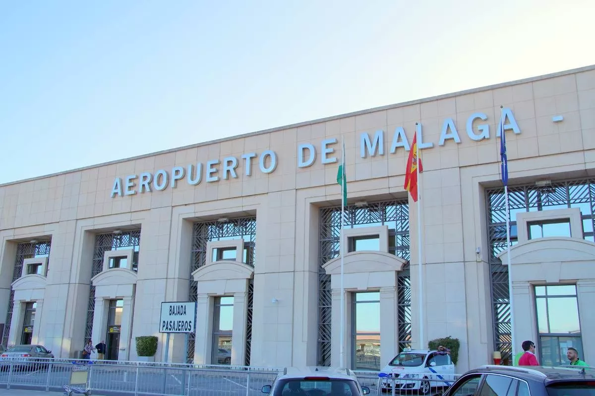 An image of a modern, large building with a sign above the entrance that reads "Universidad de Málaga" in large letters. The building has several large windows and is situated in a well-maintained area with parked cars in front. Flags are visible on the building's facade, and the sky above is clear and bright.