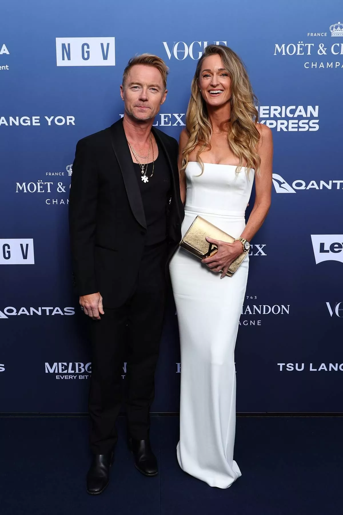 Two individuals, a man and a woman, are standing next to each other on a blue background, possibly at an event. The man is wearing a black suit and the woman is wearing a white dress. Behind them is a backdrop featuring various logos and text.
