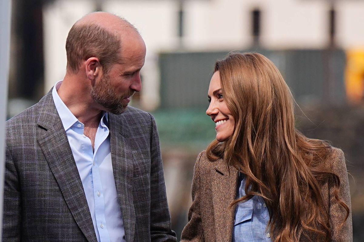 An adult male and female are standing close to each other, possibly engaged in a conversation. The man is dressed in a gray suit with a light blue shirt, while the woman wears a brown jacket and a blue top. They both appear to be smiling. Behind them, a building is visible, and the sunlight is casting shadows on the ground.