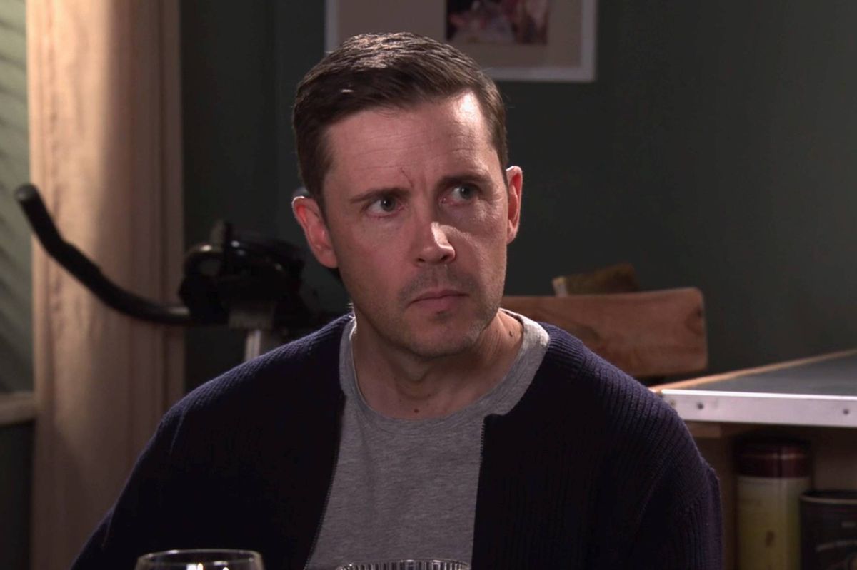 An individual is seated in an indoor setting, wearing a dark jacket over a light-colored shirt. The person's expression appears contemplative, with a slight furrow of the brow. In the background, there is a green wall adorned with a picture frame and a wooden structure. A desk with various items is partially visible in the foreground.